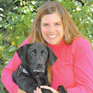 A woman with long brown hair in a pink top holds a black dog outdoors in front of green foliage, both looking at the camera.