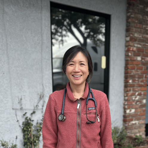 A person wearing a stethoscope and a pink fleece jacket stands smiling in front of a building with a glass door and brick wall.
