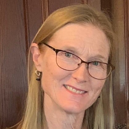 A woman with long blonde hair and glasses smiles at the camera in front of a wooden background.